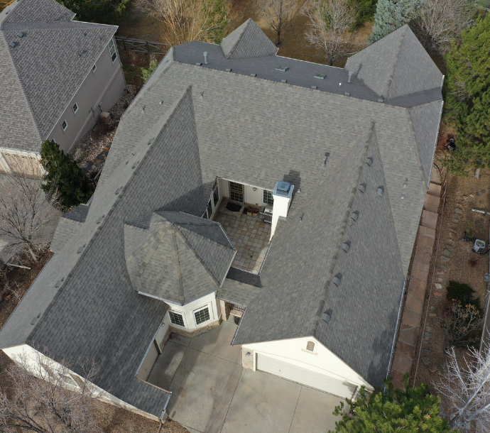 Aerial view of a large suburban home with a complex gray shingle roof, featuring various peaks and valleys, with a fenced backyard and a spacious driveway, in a residential neighborhood.