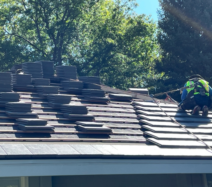 Focused roofer securing new dark slate shingles on a residential roof, with stacks of shingles ready for installation, showcasing diligence and precision in roofing work amidst a backdrop of lush greenery.
