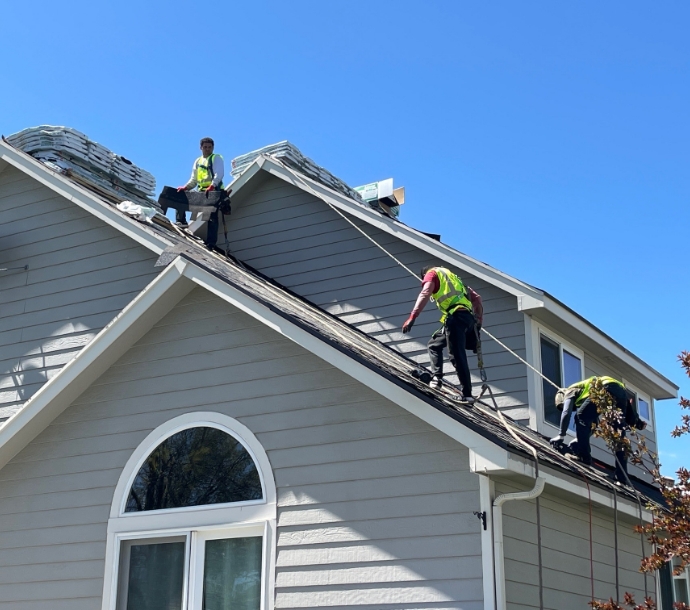 Team of roofers with high-visibility vests installing asphalt shingles on a residential roof, with safety harnesses on a sunny day, illustrating active construction and roofing safety protocols.