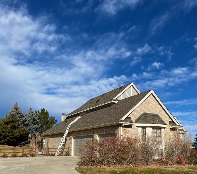 Suburban brick home with a pitched shingle roof and a ladder leaning against it under a blue sky with wispy clouds, suggesting roof maintenance on a clear day.