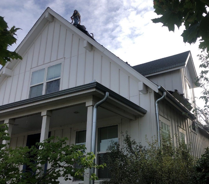 Roofer in safety gear working atop a two-story house with white siding and a black shingle roof, framed by green foliage and a clear sky, showcasing home roofing maintenance.