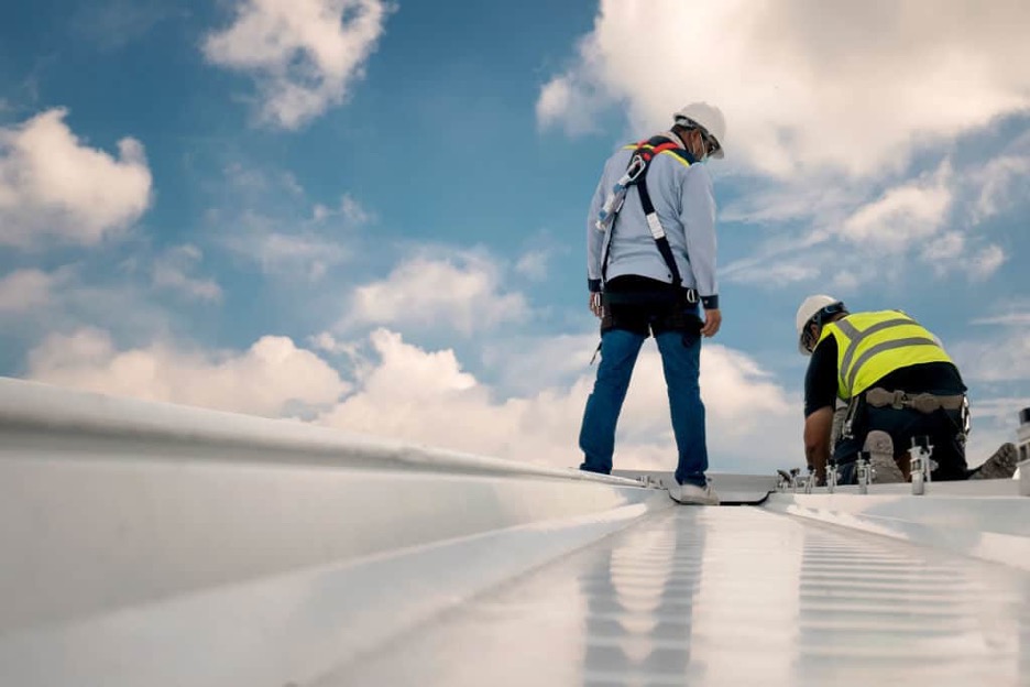 Two workers on a commercial building's white reflective TPO roof, one standing and overseeing, the other kneeling and performing installation or maintenance tasks under a partly cloudy sky.
