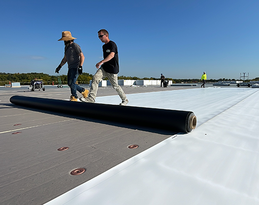 Two roofers unroll a large black EPDM rubber roofing membrane on a flat commercial roof, against a clear blue sky, demonstrating the installation process for durable synthetic roofing materials.