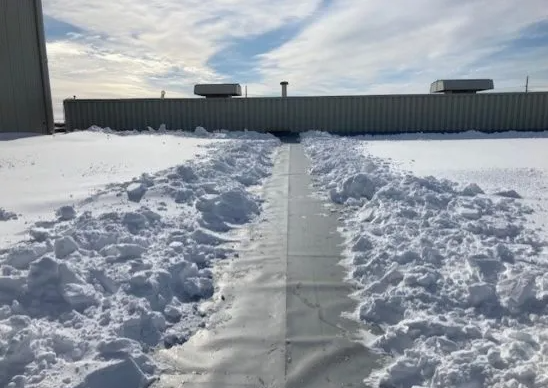 A cleared path through the snow on a flat commercial rooftop, with industrial vents and HVAC units visible against a clear winter sky, highlighting the maintenance work during snowy conditions.