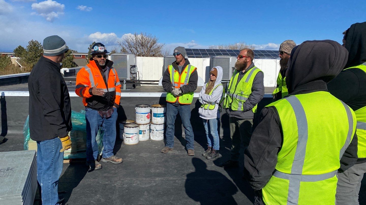 A group of construction workers in high-visibility vests engaging in a team meeting on a flat commercial rooftop, with roofing materials and equipment in the background, under a clear sky.