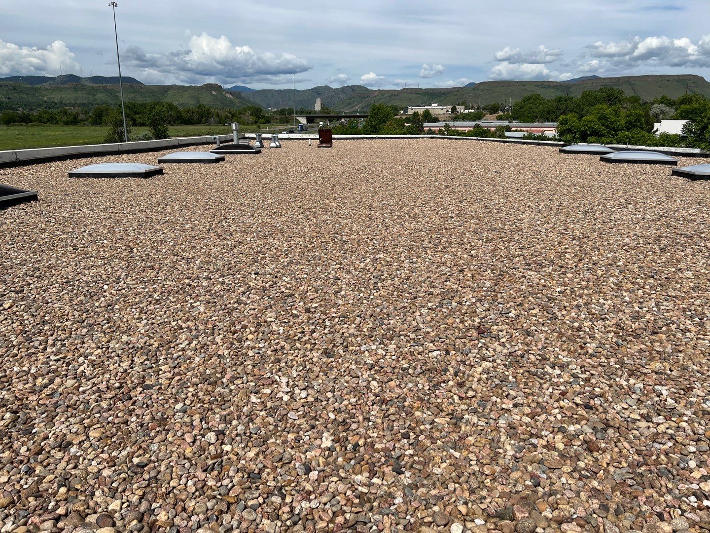 Flat commercial rooftop covered with protective gravel, with skylights and mechanical equipment, set against a backdrop of rolling hills and a cloudy sky, emphasizing the blend of functionality and natural surroundings.