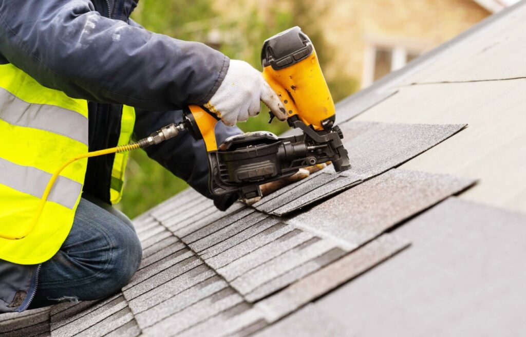 Roofer in high-visibility jacket using a pneumatic nail gun to secure shingles on a residential roof, highlighting the precision and efficiency of modern roofing techniques.