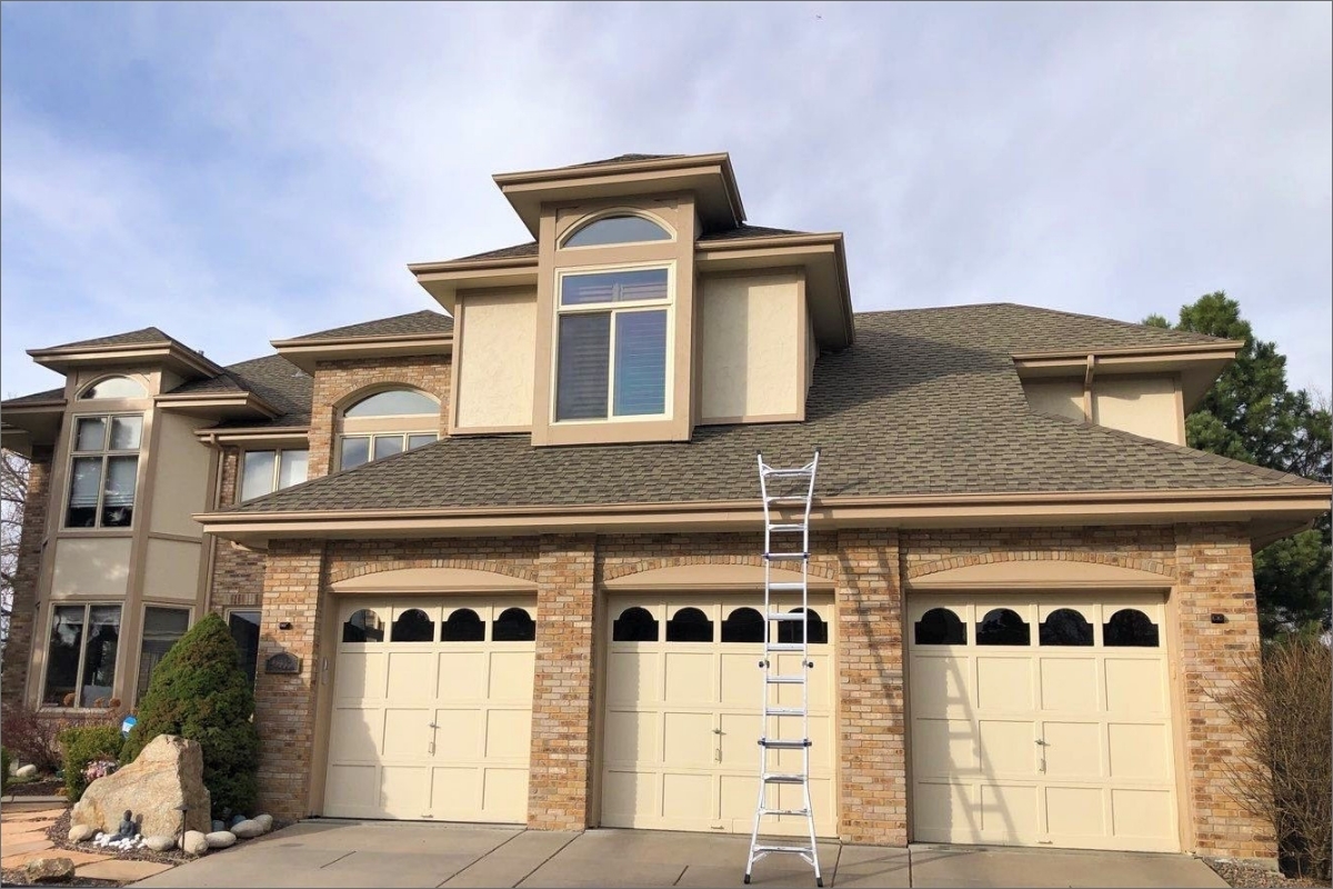 A well-maintained suburban home featuring a beige stucco exterior with stone accents, a multi-gabled dark shingle roof, and a triple garage door, with a silver extension ladder set up on the driveway for roof access.