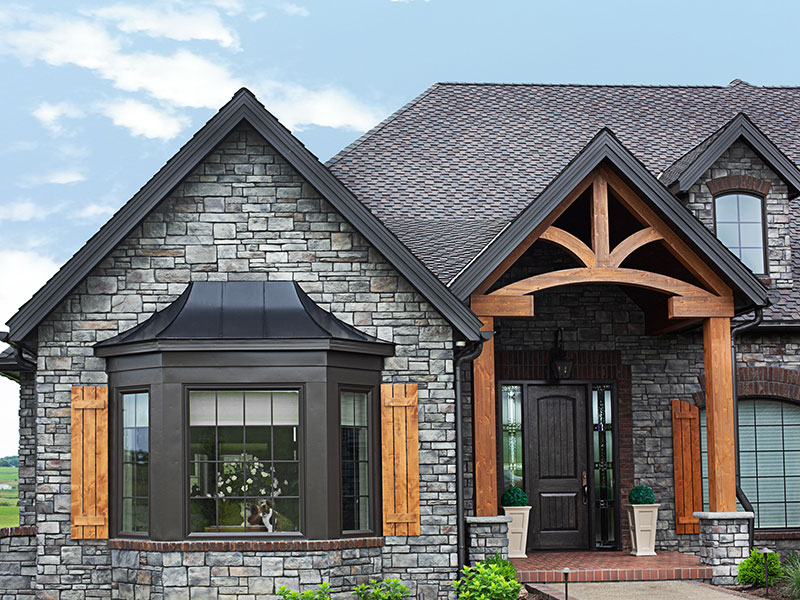 Elegant stone facade of a house with a dark shingle roof. A bay window with a copper roof and large windows sits next to a wooden front door with an arched transom and sidelights. The architectural details include wooden brackets supporting the gable over the entrance.