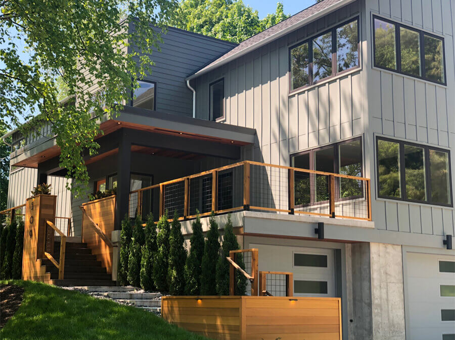 A modern multi-level home with gray and white siding, featuring large windows and wooden deck railings in a warm tone. The deck is accessible via a staircase with matching wooden handrails, and the home is surrounded by lush green trees, providing a serene residential setting.