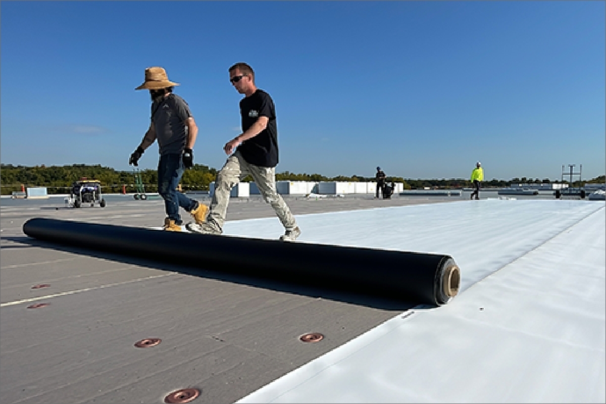wo roofers in work attire rolling out a new black EPDM rubber roofing membrane on a large commercial flat roof, under a clear blue sky, with roofing equipment visible in the background.
