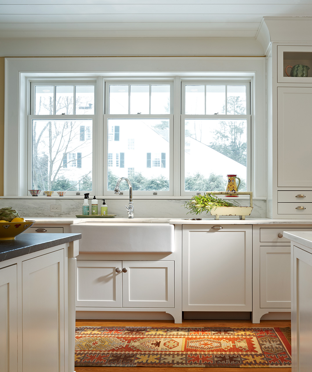 Bright kitchen interior with white cabinetry and a farmhouse sink beneath a large three-section window offering a view of a house outside. The counter is adorned with fresh fruits, herbs, and kitchen supplies. A colorful oriental rug lies on the floor, adding warmth to the space.