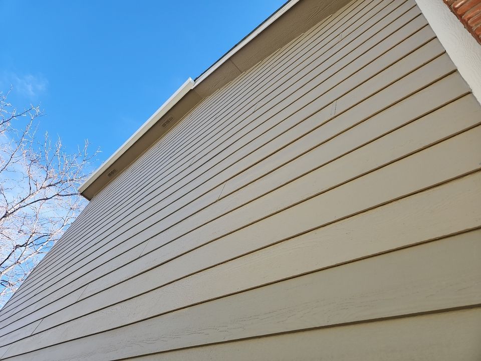 Diagonal view of a house exterior featuring cream-colored horizontal lap siding under a clear blue sky. The angle provides a view of the siding's texture and shadow lines, as well as a glimpse of bare tree branches in the upper left corner, indicating a seasonal setting.