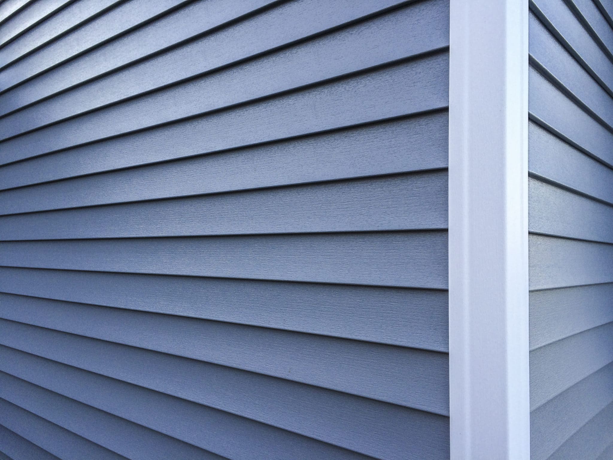 Close-up of a house corner showing blue-gray vinyl horizontal siding with a visible texture and shadow lines. A white corner post creates a crisp edge, highlighting the clean lines and durable construction of the siding material.