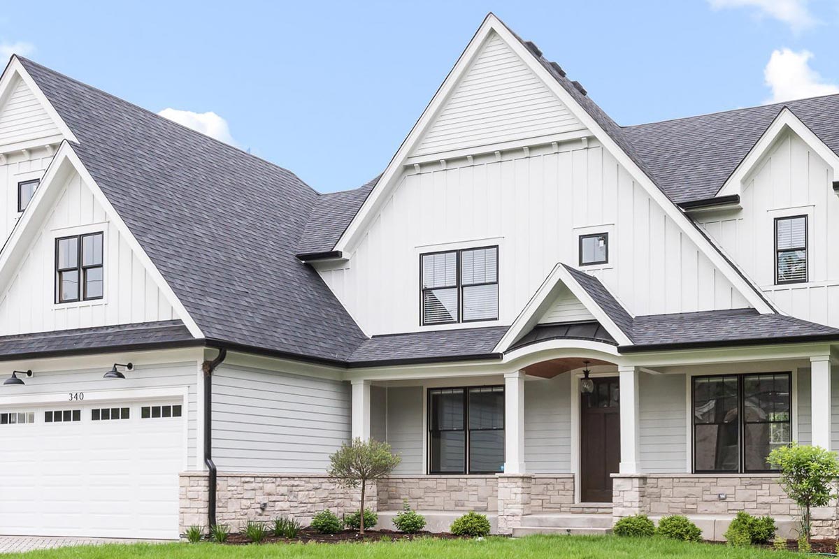 Front exterior of a luxury home with white horizontal lap siding and dark gray shingles. The architecture includes steep gable roofs, a covered porch with tapered columns over a stone base, and a double garage door. The elegant design is complemented by a neatly manicured lawn.