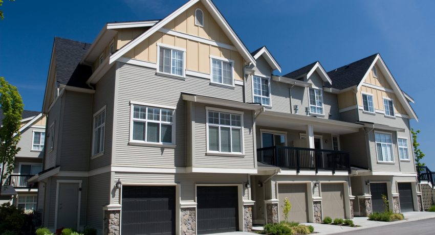 Modern multi-story townhouses with neutral color siding, stone accents, balconies, and attached garages under a clear blue sky.