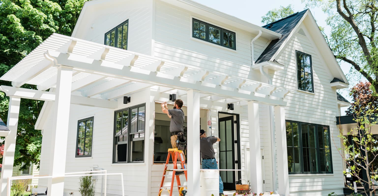 A white modern farmhouse with large windows and a covered porch supported by square columns. A person on an orange ladder is working on the porch's structure, possibly painting or installing trim. The house is surrounded by lush greenery under a bright blue sky.