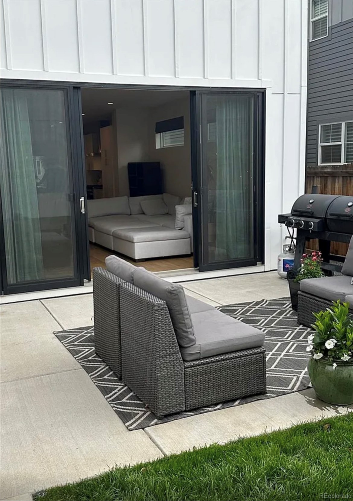 A patio area with a wicker armchair and a geometric-patterned rug. Sliding glass doors are open to an interior space with visible furniture, including a couch. The exterior siding is white, and there is a barbecue grill to the right. Lush green grass surrounds the patio.