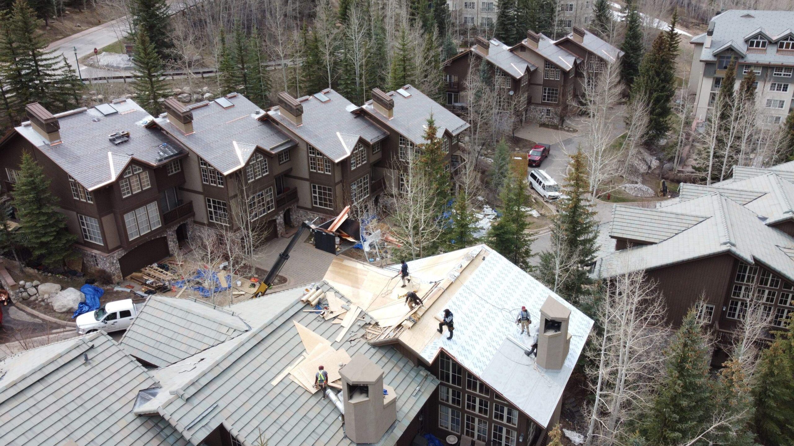 Workers on the roof of a mountain resort cabin under construction, surrounded by pine trees and adjacent cabins with snow on the ground.