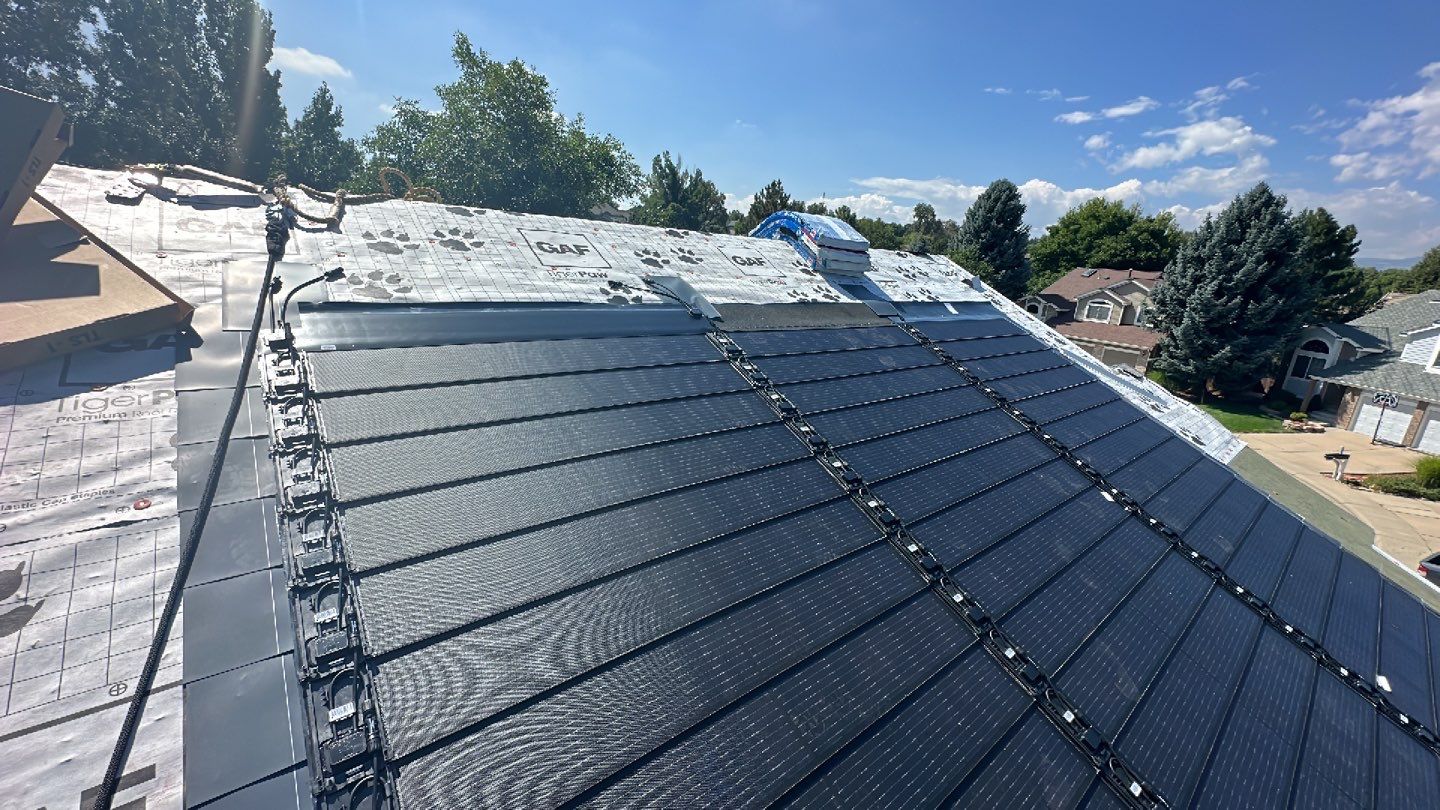 Roof with dark shingles and solar panels against a blue sky.