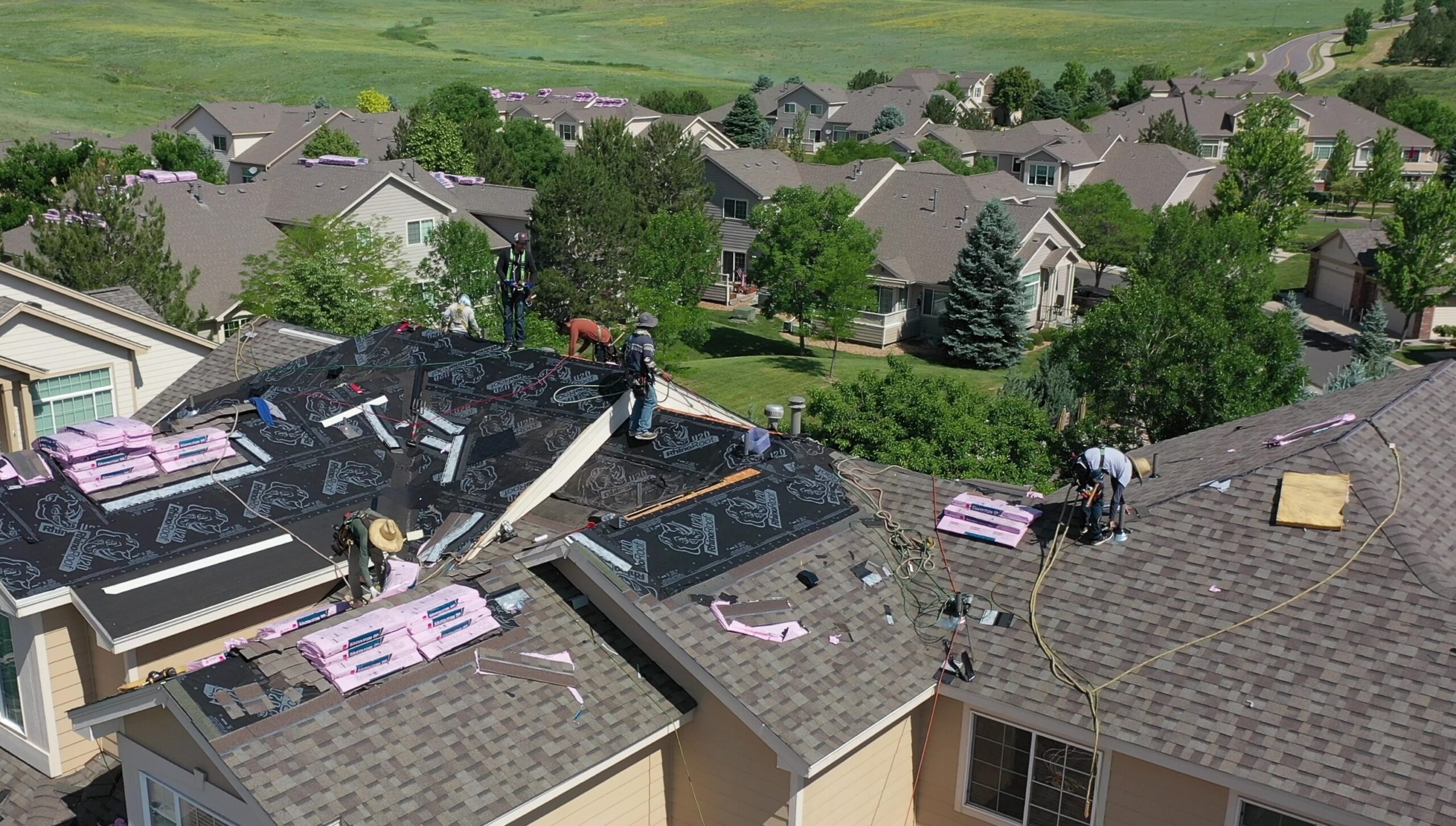 Roofers at work on a residential building with new roofing material and packs of Owens Corning insulation shingles visible, in a suburban neighborhood.