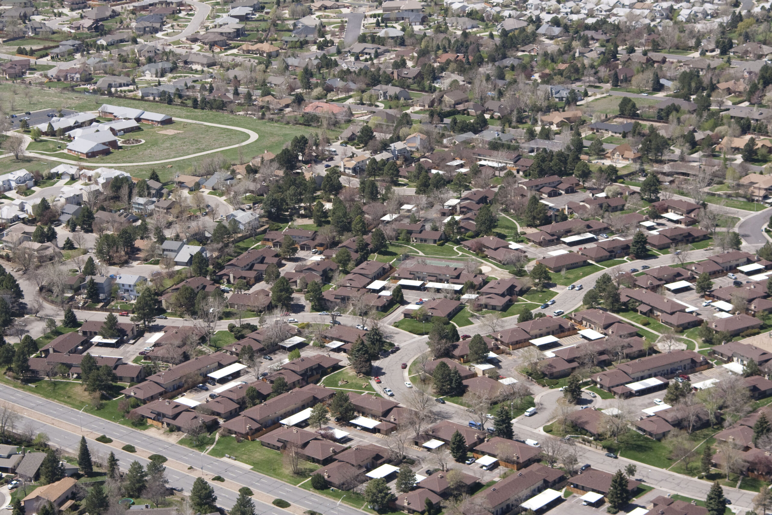 The image depicts a residential area near Denver, Colorado, with a view from above that showcases the houses and streets, greenery, and a clear view of the surrounding environment. The area is characterized by a number of homes with similar roofing, likely indicating a suburban neighborhood with well-organized streets and landscaping.