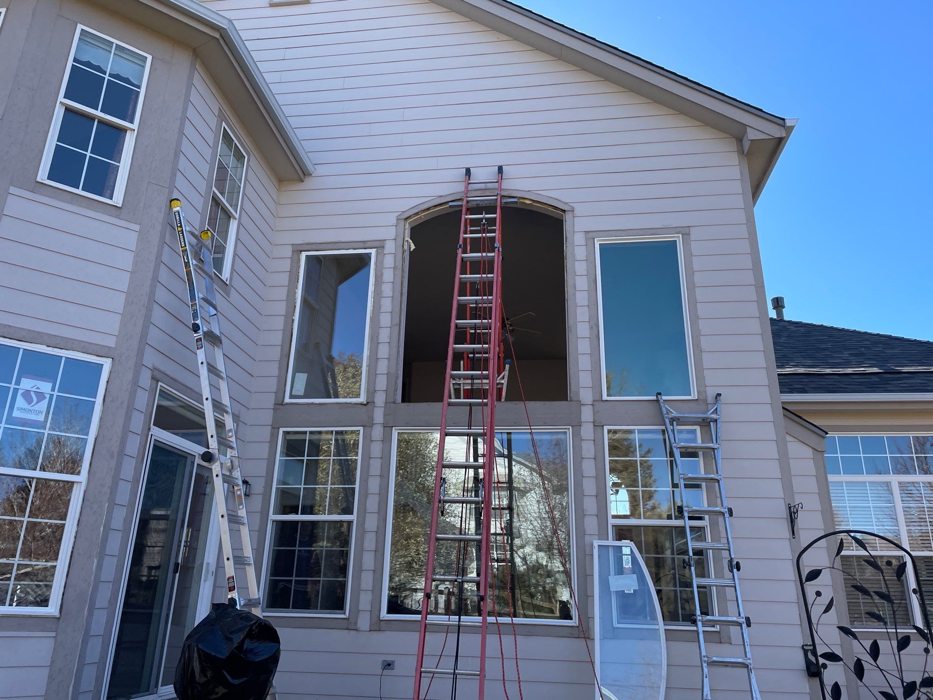 Exterior view of a two-story house with a beige siding finish and multiple large windows. There is a red extension ladder and a silver step ladder set against the house, presumably for maintenance work on the upper windows. A blue sky with a few clouds is visible above the house.