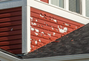 A close-up of a building's exterior shows red siding with extensive hail damage, exposing the underlying white surface. siding damage insurance claim