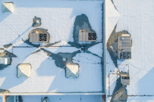 A commercial roof covered in snow, with a few vents and chimneys visible. Scene is calm and peaceful, as the snow covers the roof and the vents and chimneys are barely visible